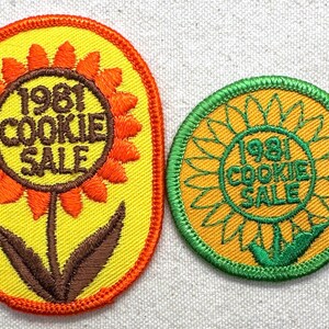 May include: Two vintage embroidered patches. One is oval, yellow with an orange sunflower design and the text "1981 COOKIE SALE". The other is round, yellow with a green sunflower design and the same text.