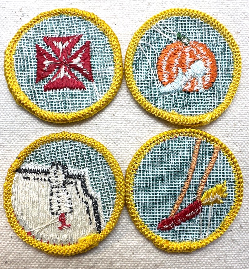 May include: Four round embroidered patches with yellow borders. The patches feature designs of a red cross, an orange pumpkin, a white and black figure, and a red and yellow object. The patches are on a light green mesh background.