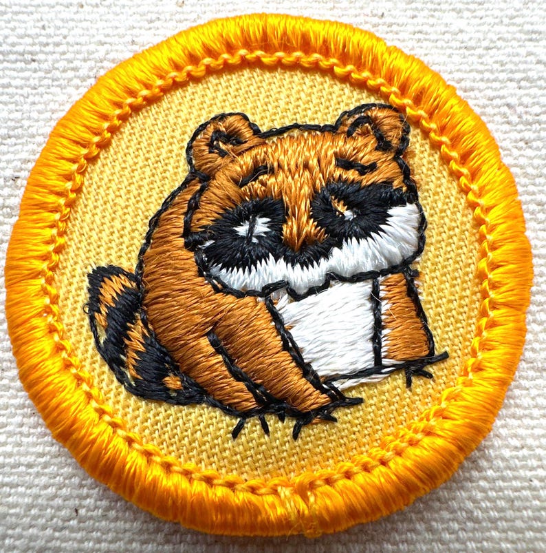 May include: Embroidered patch featuring a cartoon raccoon in shades of brown, black, and white. The raccoon is centered on a yellow background with a yellow stitched border. The patch is round and has a textured appearance.