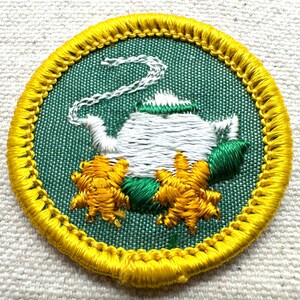 May include: Embroidered patch featuring a white teapot with a green lid and steam, surrounded by yellow and green floral accents. The patch has a green background and a yellow border. The design is circular.
