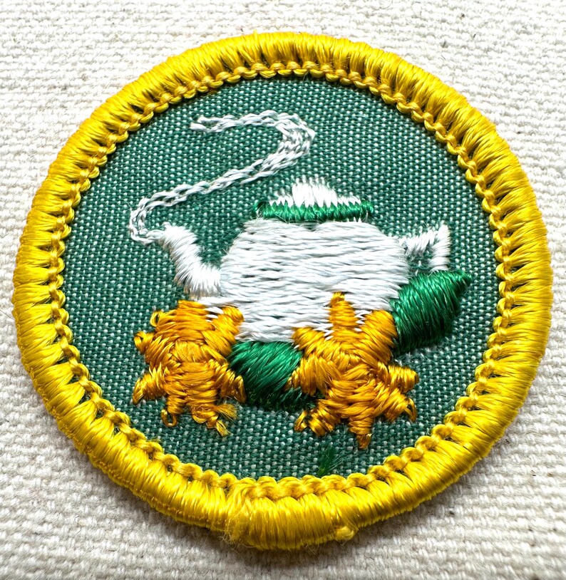 May include: Embroidered patch featuring a white teapot with a green lid and steam, surrounded by yellow and green floral accents. The patch has a green background and a yellow border. The design is circular.