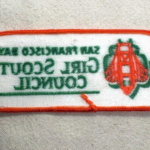 May include: A rectangular patch with an orange border and white background. The patch features the text "SAN FRANCISCO BAY GIRL SCOUT COUNCIL" in green. An orange illustration of the Golden Gate Bridge is on the right side.
