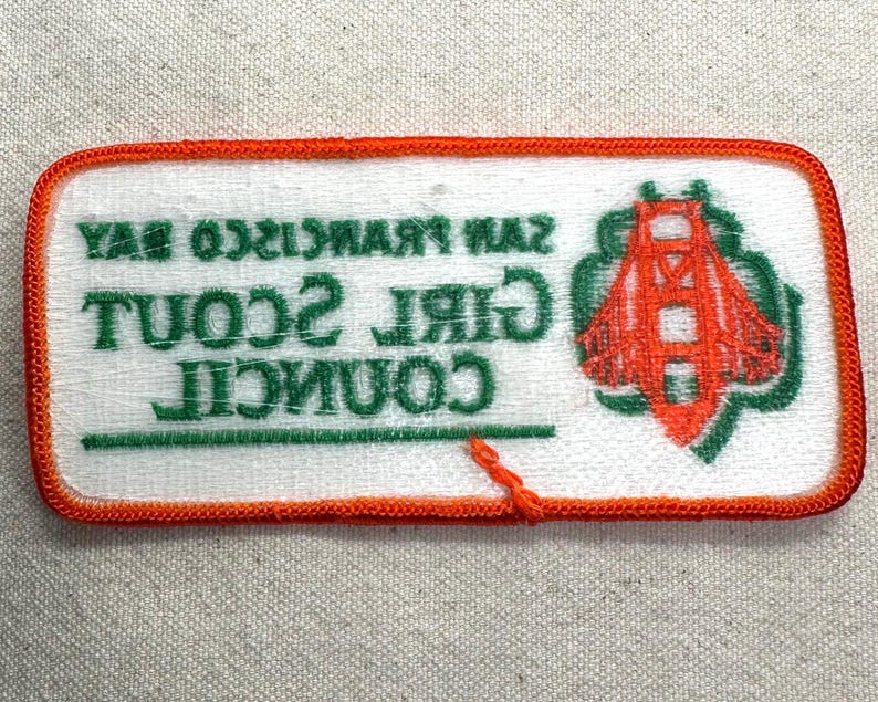May include: A rectangular patch with an orange border and white background. The patch features the text "SAN FRANCISCO BAY GIRL SCOUT COUNCIL" in green. An orange illustration of the Golden Gate Bridge is on the right side.