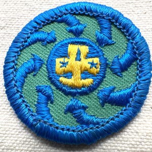 May include: Circular embroidered patch with a blue border and a green center. The center features a blue circle with a yellow cross design. Blue arrows encircle the center, creating a dynamic visual.