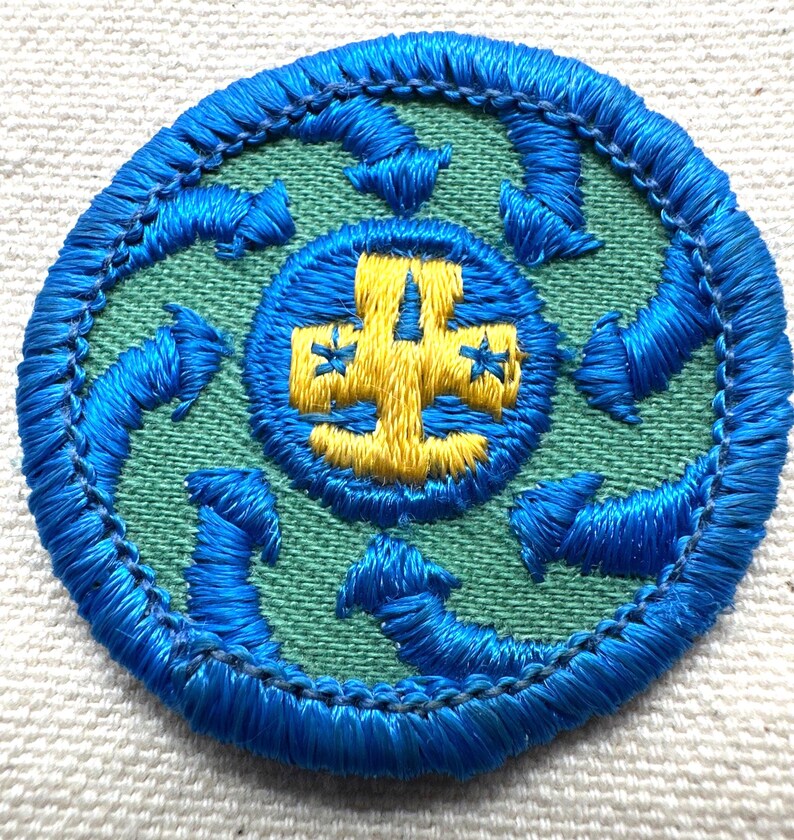 May include: Circular embroidered patch with a blue border and a green center. The center features a blue circle with a yellow cross design. Blue arrows encircle the center, creating a dynamic visual.