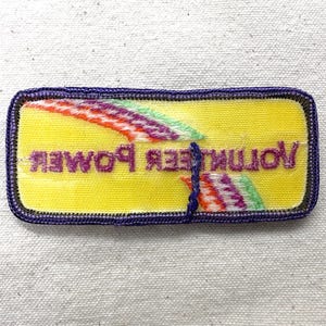 May include: A rectangular patch with a yellow background and the words "Volunteer Power" in purple. The patch has a purple border and colorful diagonal stripes.