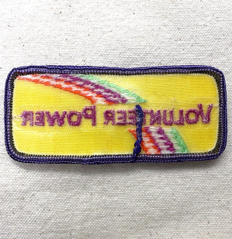 May include: A rectangular patch with a yellow background and the words "Volunteer Power" in purple. The patch has a purple border and colorful diagonal stripes.