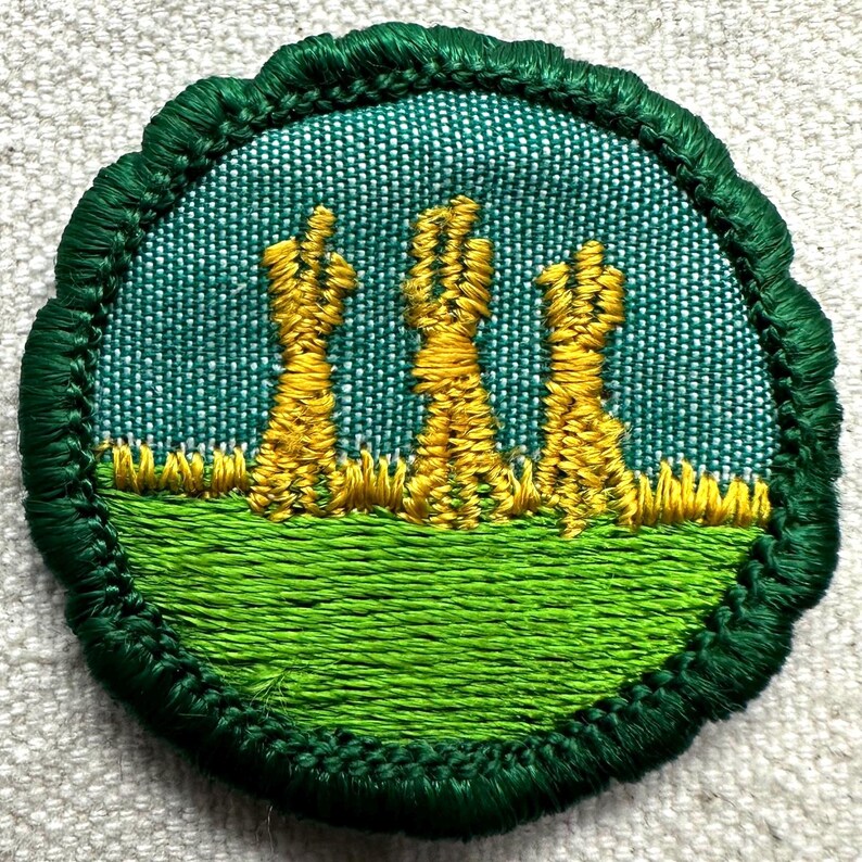 May include: Embroidered patch featuring a green border and a central design. The design depicts three yellow wheat sheaves against a teal background, above a green field. The patch is round with a scalloped edge.