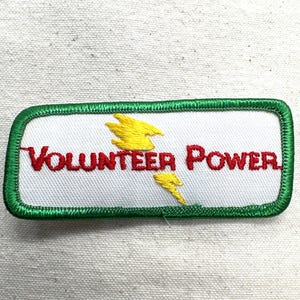May include: Embroidered patch with the words "VOLUNTEER POWER" in red, with yellow lightning bolt graphics. The patch has a white background and a green border.