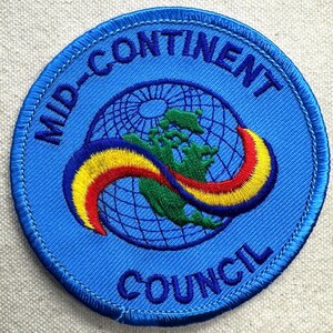May include: A circular embroidered patch with a blue background featuring a globe graphic in the center. The text MID-CONTINENT is arched across the top and COUNCIL is arched across the bottom in blue. A curved rainbow ribbon crosses the globe.