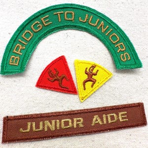 May include: A collection of embroidered patches. A green arched patch reads "BRIDGE TO JUNIORS" in gold thread. Two triangular patches, one red and one yellow, feature a brown figure. A brown rectangular patch reads "JUNIOR AIDE" in gold.