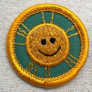 Puede incluir: Parche bordado con un sol sonriente con contorno y rayos amarillos, sobre un fondo verde azulado. El sol tiene ojos cuadrados negros y una sonrisa negra curvada. El parche es circular y tiene un borde amarillo.