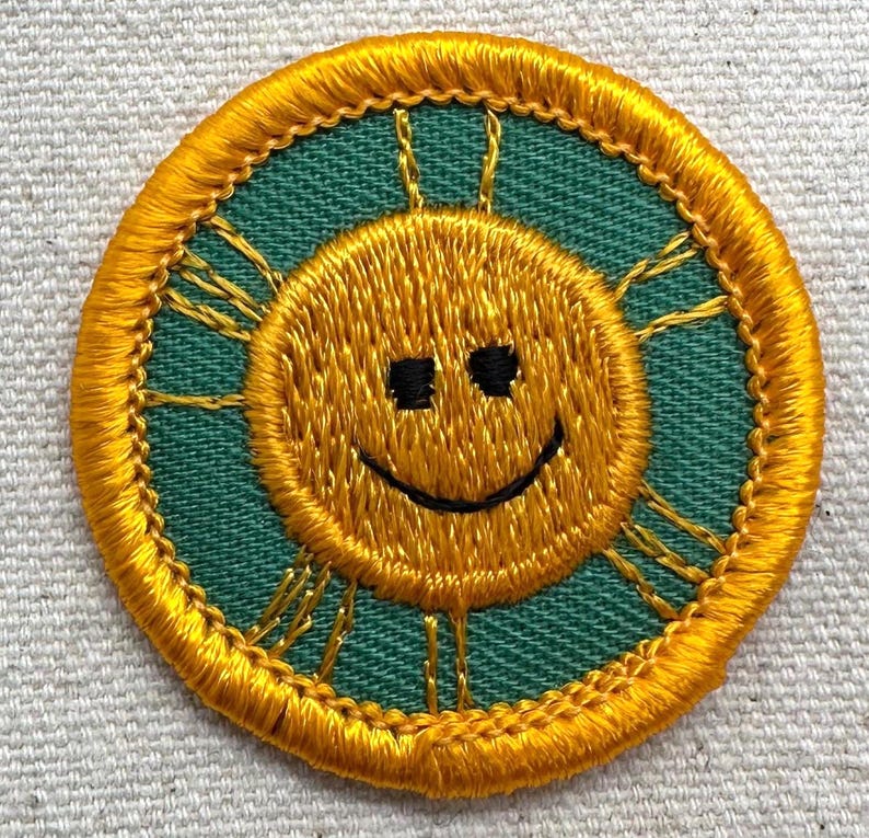 Puede incluir: Parche bordado con un sol sonriente con contorno y rayos amarillos, sobre un fondo verde azulado. El sol tiene ojos cuadrados negros y una sonrisa negra curvada. El parche es circular y tiene un borde amarillo.