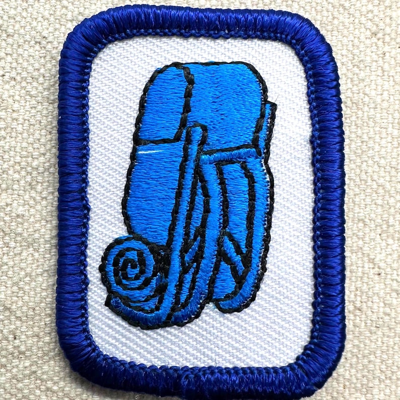 May include: Embroidered patch featuring a blue backpack and rolled sleeping bag design. The patch has a white background and a blue border. The backpack and sleeping bag are detailed with black stitching.