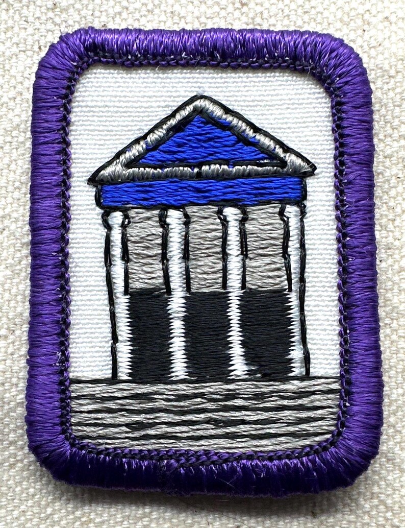 May include: Embroidered patch featuring a building with a blue roof and columns. The patch has a white background and a purple border. The building's columns are black, white, and gray. The patch is rectangular.