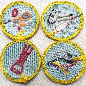 May include: Four round embroidered patches with yellow stitched borders. Each patch features a different design: a lacrosse stick, a rocket, a red tool, and a bow and arrow. The patches have a light blue background.