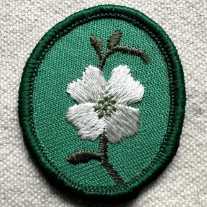 May include: Embroidered patch featuring a white flower with green center and stem, set against a green oval background. The patch is bordered with a dark green stitched edge, creating a textured appearance. The flower's petals are detailed with white thread.
