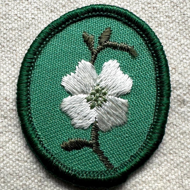 Op de afbeelding: Geborduurde patch met een witte bloem en groene stengel op een groen ovaal. De patch heeft een donkergroene gestikte rand. De bloemblaadjes zijn gedetailleerd met witte draad. Geschikt voor kleding of accessoires.