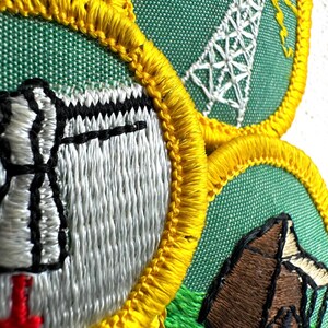 May include: Close-up of three embroidered patches with green backgrounds and yellow borders. The patches feature images of a radio tower, a tent, and a stylized object. The embroidery is in white, black, red, and brown.