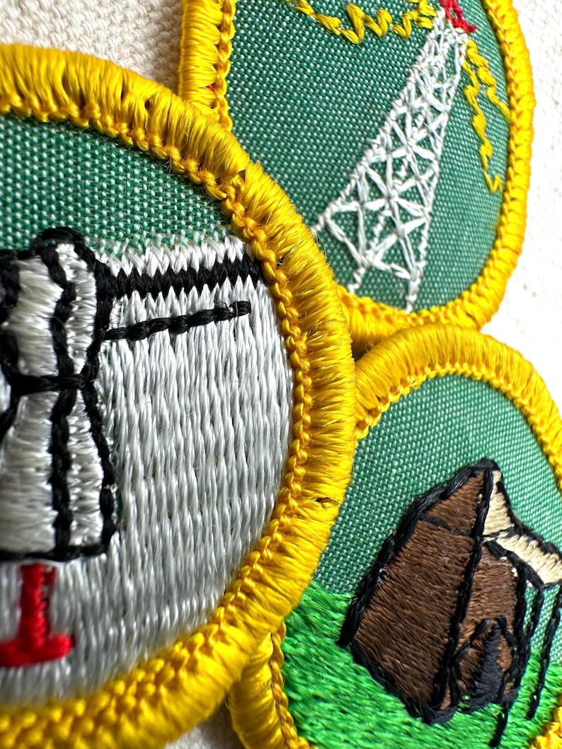 May include: Close-up of three embroidered patches with green backgrounds and yellow borders. The patches feature images of a radio tower, a tent, and a stylized object. The embroidery is in white, black, red, and brown.