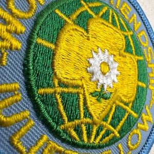 Peut inclure: &Eacute;cusson brod&eacute; avec les mots "WORLD FRIENDSHIP" et "JULIETTE LOW" en fil jaune. Le patch pr&eacute;sente un motif de globe vert et jaune avec une fleur blanche au centre, le tout sur un fond bleu clair.