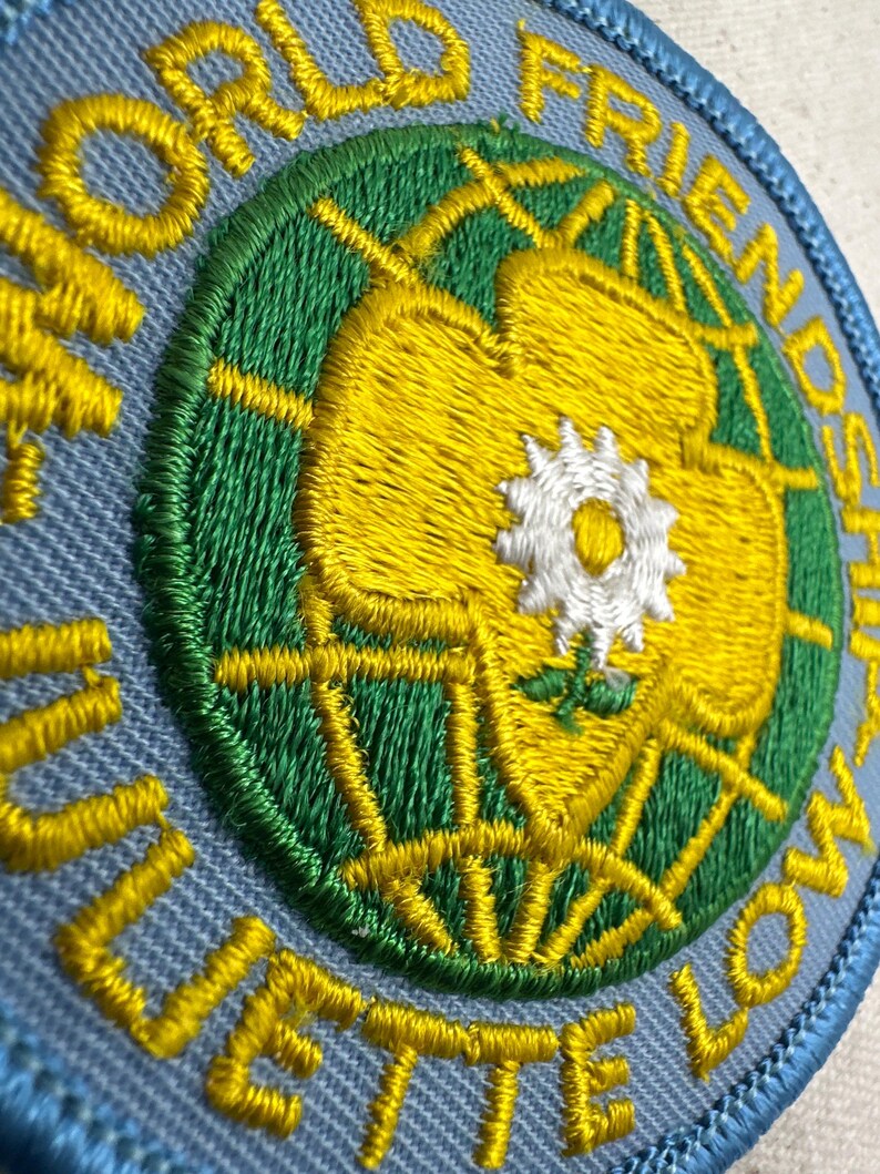 Peut inclure: &Eacute;cusson brod&eacute; avec les mots "WORLD FRIENDSHIP" et "JULIETTE LOW" en fil jaune. Le patch pr&eacute;sente un motif de globe vert et jaune avec une fleur blanche au centre, le tout sur un fond bleu clair.