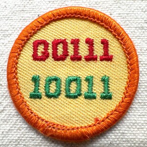 May include: A round patch with an orange border and a yellow center. The top text reads "00111" in red, and the bottom text reads "10011" in green. The patch is likely for a club or organization.