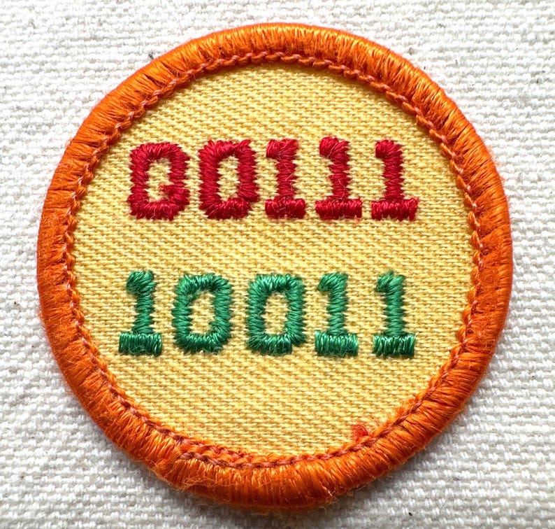 May include: A round patch with an orange border and a yellow center. The top text reads "00111" in red, and the bottom text reads "10011" in green. The patch is likely for a club or organization.