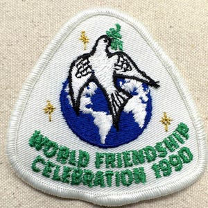 May include: Embroidered patch with a white dove over a blue globe, with the text "WORLD FRIENDSHIP CELEBRATION 1990" in green. The patch has a white border and gold star accents. The patch is triangular in shape.