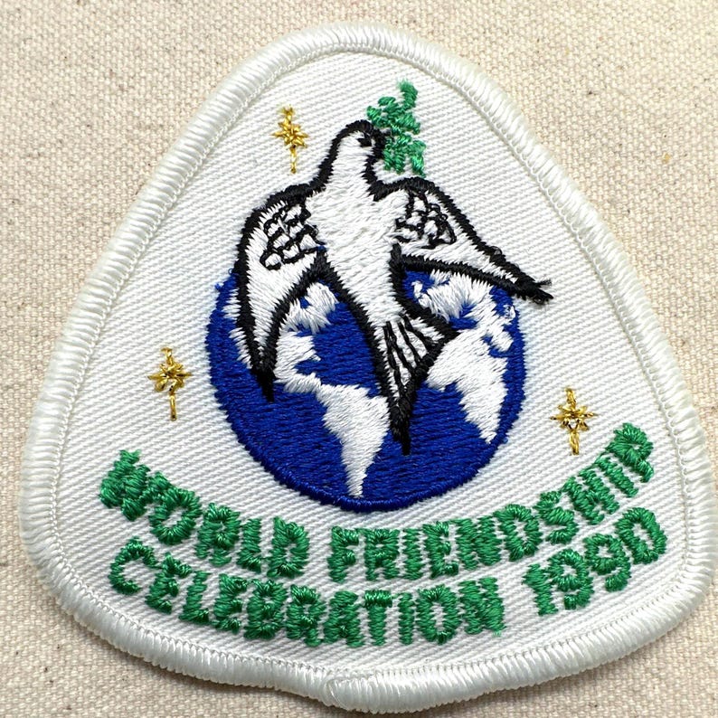 May include: Embroidered patch with a white dove over a blue globe, with the text "WORLD FRIENDSHIP CELEBRATION 1990" in green. The patch has a white border and gold star accents. The patch is triangular in shape.