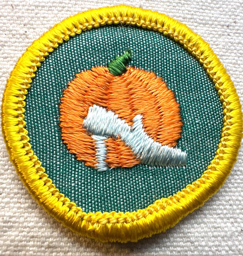 May include: Embroidered patch featuring an orange pumpkin with a green stem and a light blue letter "Q" on a green background. The patch is round with a yellow border.