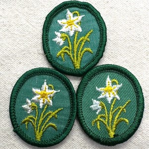May include: Three embroidered patches with a green oval border. Each patch features a white and yellow flower design with green stems and leaves on a green background. The patches are likely for clothing or crafts.