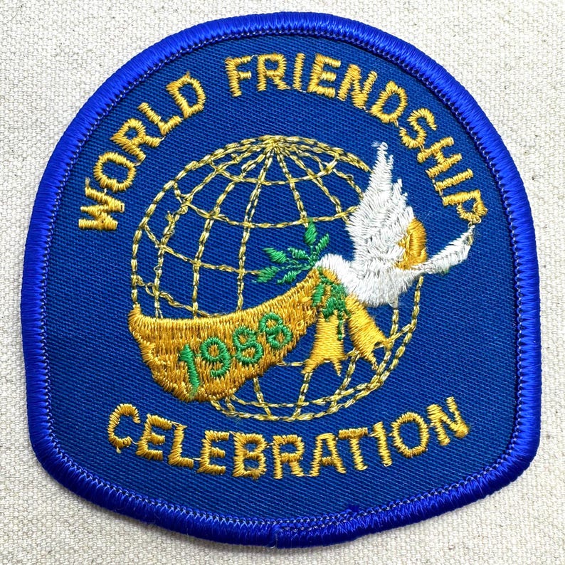 K&ouml;nnte beinhalten: Blauer, gestickter Aufn&auml;her mit den Worten "WORLD FRIENDSHIP CELEBRATION" und "1998". Der Aufn&auml;her zeigt einen goldenen Globus, eine wei&szlig;e Taube und ein Banner mit gr&uuml;nen Bl&auml;ttern.