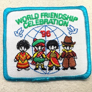 May include: Embroidered patch with a white background and a blue border. The patch features the text "WORLD FRIENDSHIP CELEBRATION '96" above a globe graphic and four cartoon figures in colorful outfits. Two doves are on either side of the globe.