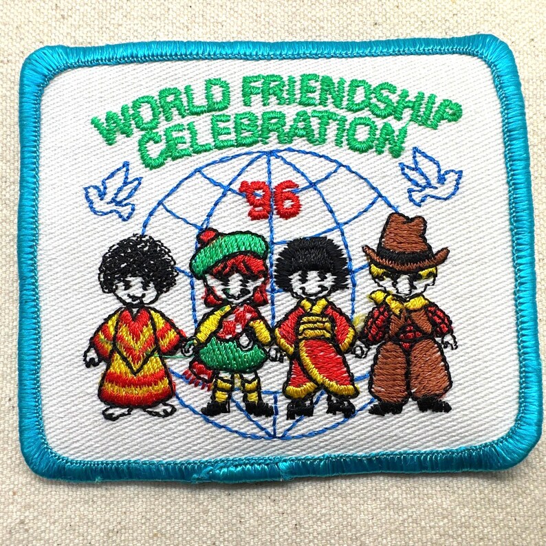 May include: Embroidered patch with a white background and a blue border. The patch features the text "WORLD FRIENDSHIP CELEBRATION '96" above a globe graphic and four cartoon figures in colorful outfits. Two doves are on either side of the globe.
