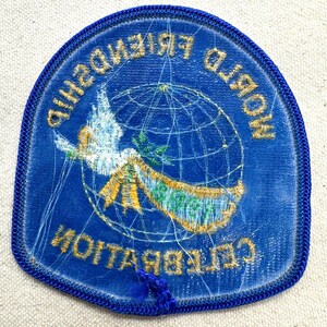 K&ouml;nnte beinhalten: Blauer, gestickter Aufn&auml;her mit den Worten "WORLD FRIENDSHIP CELEBRATION" in goldfarbenen Buchstaben. Der Aufn&auml;her zeigt eine wei&szlig;e Taube, die ein Banner &uuml;ber einem Globus tr&auml;gt. Der Aufn&auml;her hat einen blauen Rand.