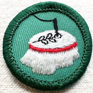 May include: Embroidered patch with a green border and background. The patch features a white and red design with a black stem and a sprig of leaves. The patch is circular and has a stitched edge.