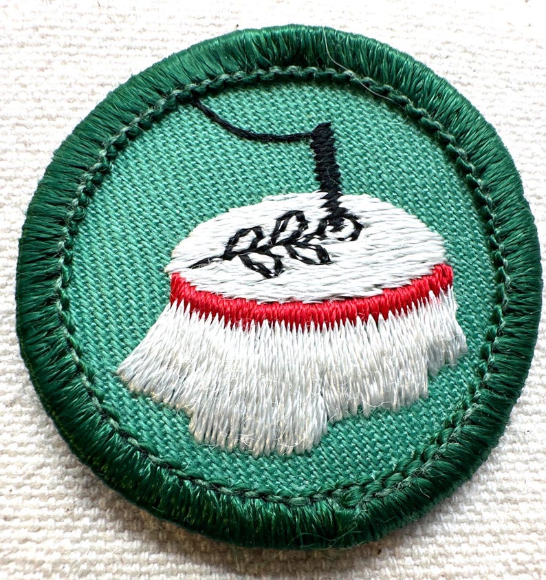 May include: Embroidered patch with a green border and background. The patch features a white and red design with a black stem and a sprig of leaves. The patch is circular and has a stitched edge.