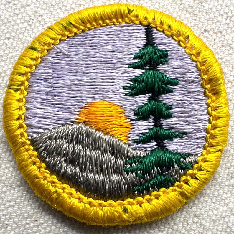May include: Embroidered patch featuring a landscape scene. A yellow sun sets behind a gray mountain, with a green pine tree on the right. The scene is set against a lavender sky, all within a yellow border.