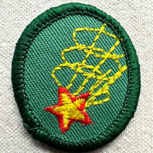 May include: An oval-shaped green embroidered patch with a yellow and red starburst design. The star is red with yellow accents, and the starburst trails are yellow. The patch has a green border.