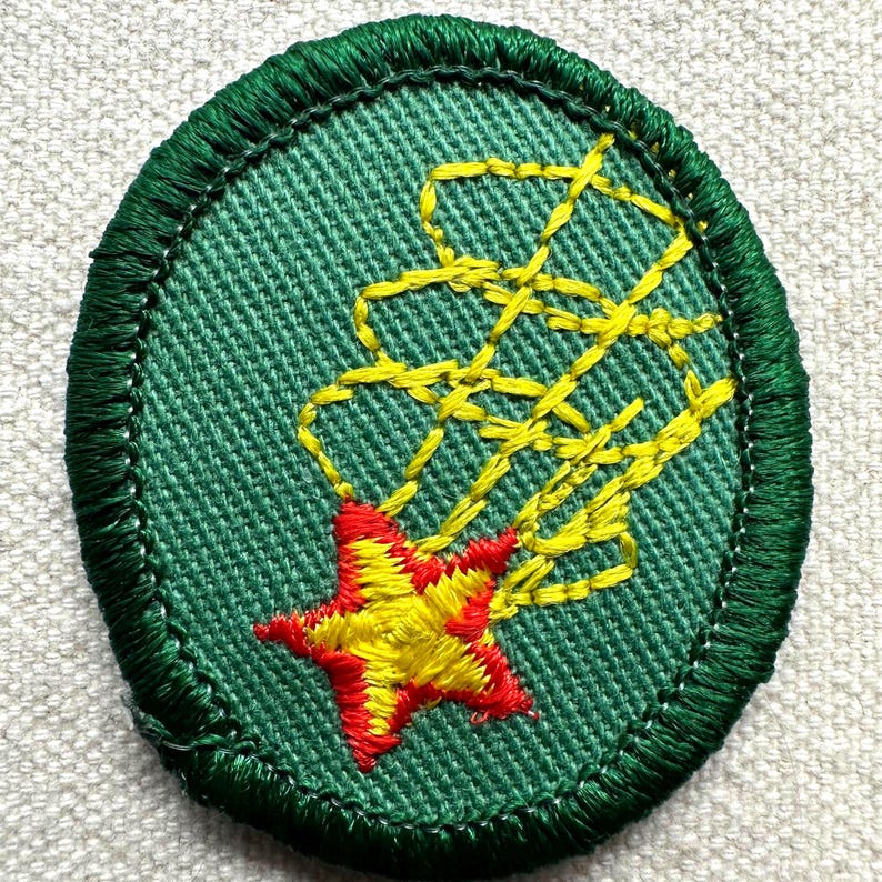 May include: An oval-shaped green embroidered patch with a yellow and red starburst design. The star is red with yellow accents, and the starburst trails are yellow. The patch has a green border.