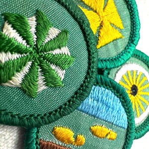 May include: Close-up of several embroidered patches with green, white, yellow, and blue designs. The patches have a green border and feature various motifs, including a starburst, a cross, a landscape, and a sun-like design. The embroidery is detailed and colorful.