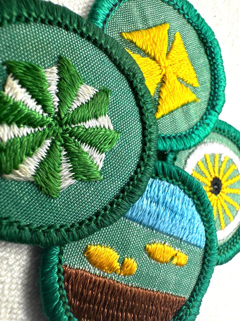 May include: Close-up of several embroidered patches with green, white, yellow, and blue designs. The patches have a green border and feature various motifs, including a starburst, a cross, a landscape, and a sun-like design. The embroidery is detailed and colorful.