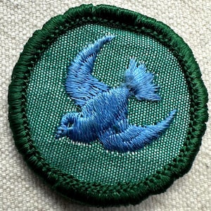 May include: Round embroidered patch featuring a blue bird in flight against a green background. The bird is intricately stitched, with a detailed wing design. The patch has a dark green border.