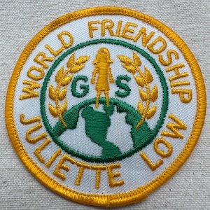 May include: Embroidered patch with the words "WORLD FRIENDSHIP JULIETTE LOW" in gold lettering. The patch features a green and gold design with a globe, a silhouette of a person, and the letters "G S".