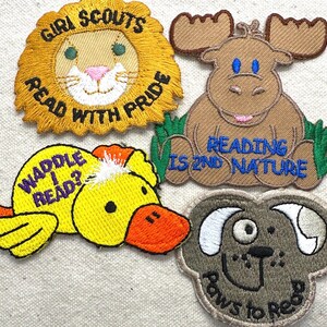 May include: Four embroidered patches with reading-themed designs. Includes a lion with "Girl Scouts Read With Pride", a moose with "Reading is 2nd Nature", a duck with "Waddle I Read?", and a dog with "Paws to Read".