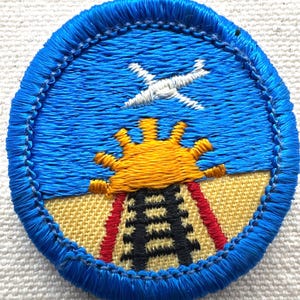 May include: Embroidered patch with a blue border, featuring a blue sky with a white airplane, a yellow sun, and a black and red structure. The patch has a textured appearance.