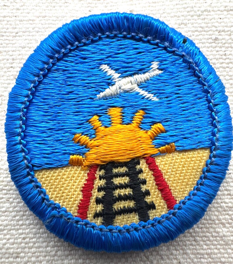 May include: Embroidered patch with a blue border, featuring a blue sky with a white airplane, a yellow sun, and a black and red structure. The patch has a textured appearance.