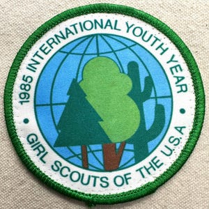 Op de afbeelding: Een ronde patch met een groene rand en witte tekst. De tekst luidt "1985 INTERNATIONAL YOUTH YEAR GIRL SCOUTS OF THE U.S.A." Het midden toont een wereldbol, een boom en een cactus in groen en blauw.