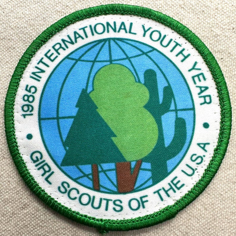 Op de afbeelding: Een ronde patch met een groene rand en witte tekst. De tekst luidt "1985 INTERNATIONAL YOUTH YEAR GIRL SCOUTS OF THE U.S.A." Het midden toont een wereldbol, een boom en een cactus in groen en blauw.